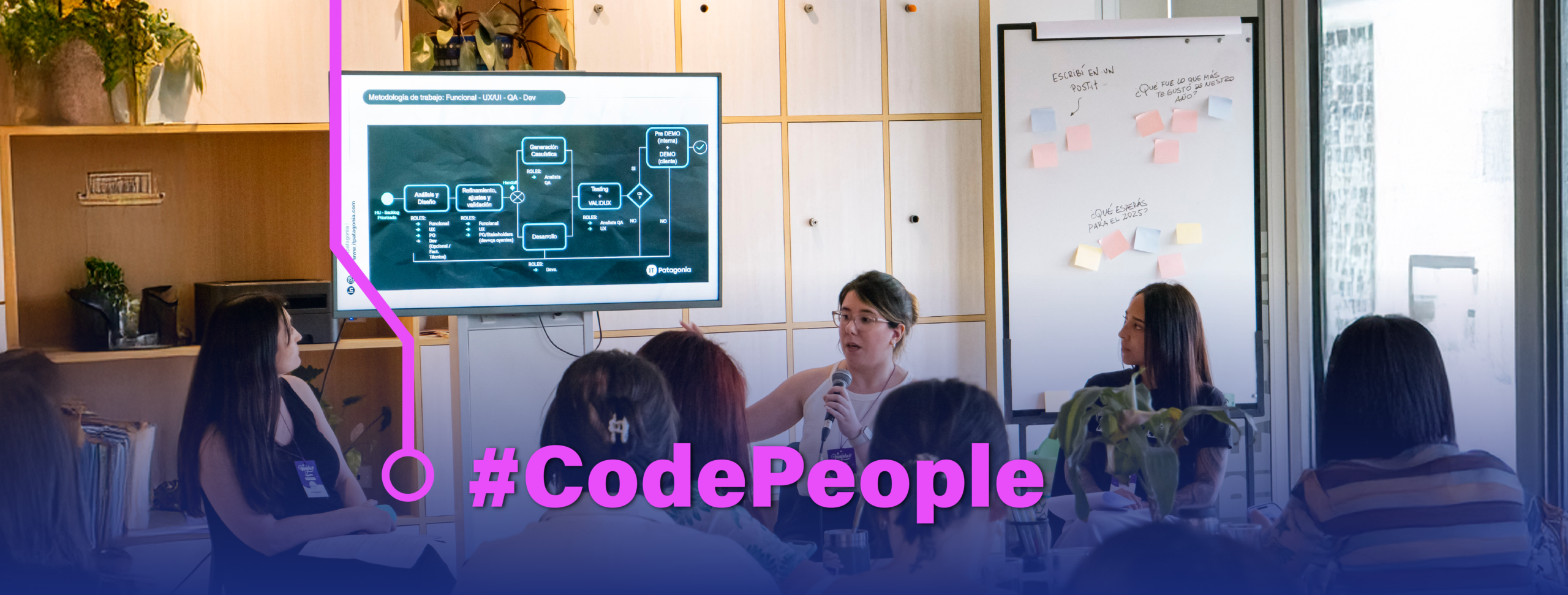 En la imagen se ve a un grupo de personas jóvenes reunidas en una formación sobre tecnología. Una mujer joven está compartiendo información a una audiencia.